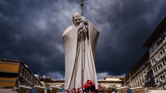 Velas y flores junto a la estatua de Juan Pablo II a las puertas del hospital Gemelli, donde el Papa Francisco está ingresado por una neumonía, en Roma, el 27 de febrero de 2025. (DIMITAR DILKOFF / AFP)